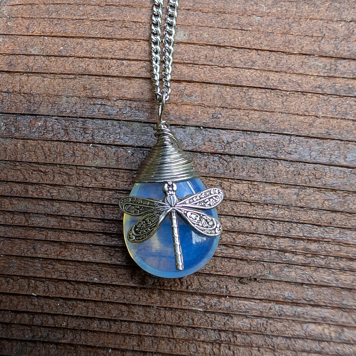 A silver dragonfly pendant necklace resting on a wooden surface. The pendant features a blue synthetic opal stone with a detailed dragonfly.