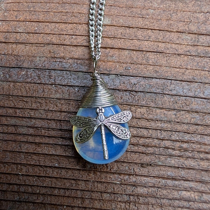 A silver dragonfly pendant necklace resting on a wooden surface. The pendant features a blue synthetic opal stone with a detailed dragonfly.