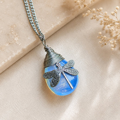 A silver dragonfly pendant necklace resting on a light beige textured surface with dried flowers. The pendant features a blue synthetic opal stone  with a detailed dragonfly.