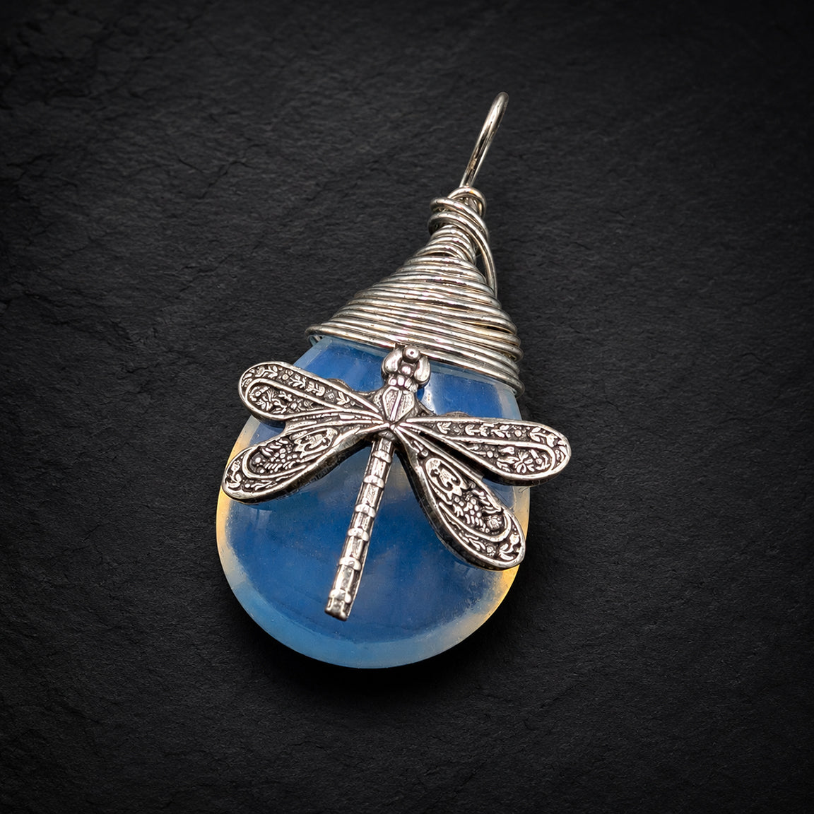 A silver dragonfly pendant necklace resting on a black background. The pendant features a blue synthetic opal stone with a detailed dragonfly.