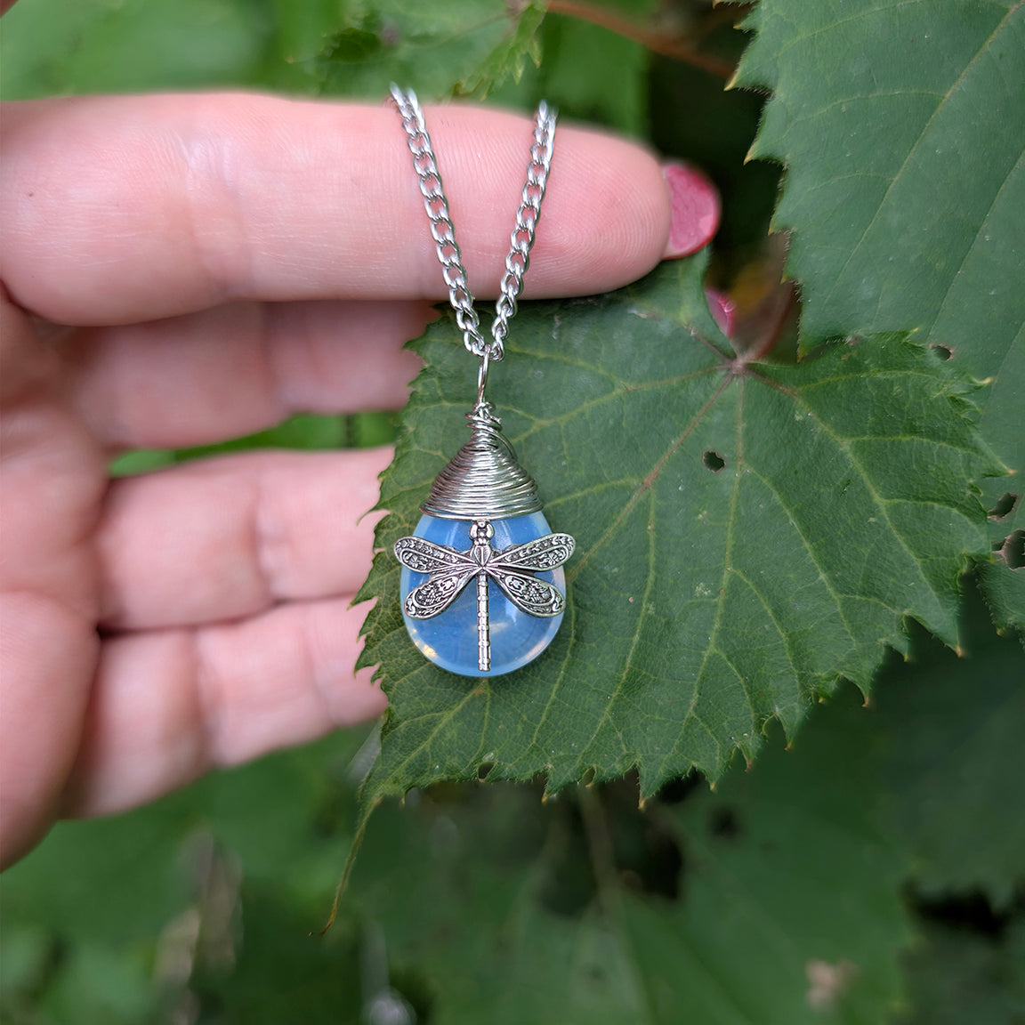 A silver dragonfly pendant with a blue stone, held by a hand against a background of green leaves.