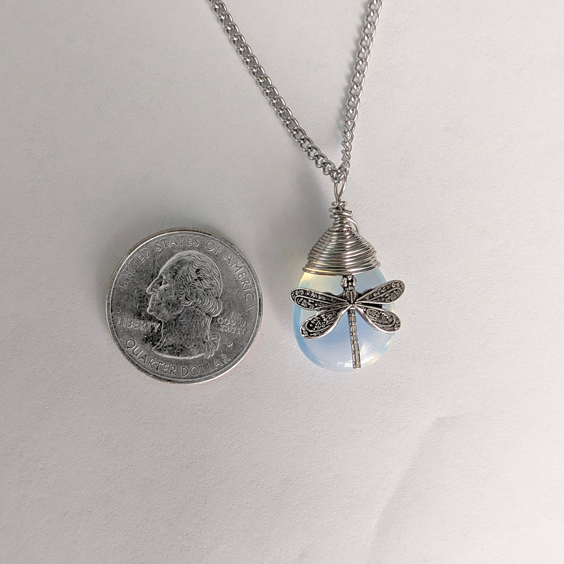 A silver dragonfly pendant necklace resting on a plain white background with a quarter for size scale. The pendant features a blue synthetic opal stone with a detailed dragonfly.