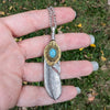 Feather-shaped pendant with turquoise stone held in a hand against a grassy background