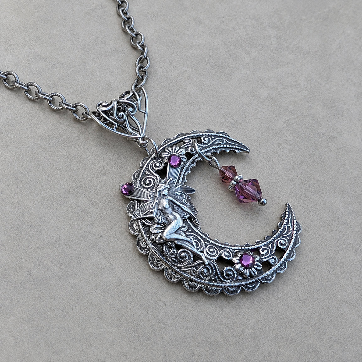 An ornate silver crescent moon pendant necklace featuring a fairy figure, small purple crystal flowers, and dangling purple crystal beads, displayed on a neutral textured beige background.
