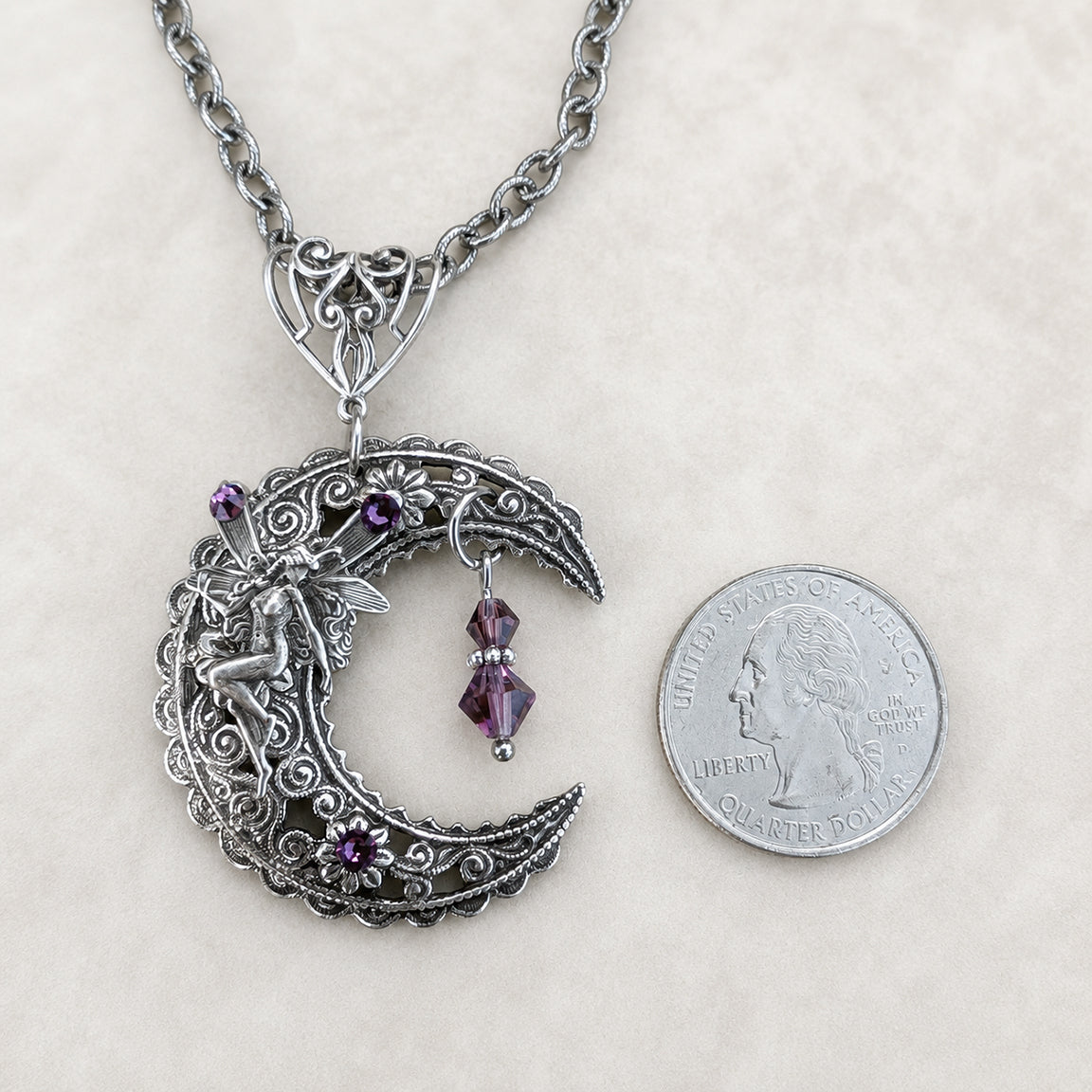 An ornate silver crescent moon pendant necklace with a fairy figure, purple gemstone accents, and a dangling purple bead, displayed next to a US quarter coin for scale on a light beige textured background.