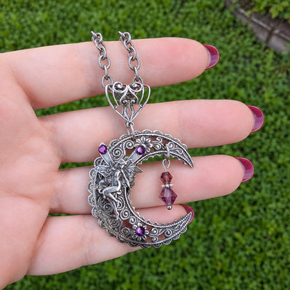 A hand with dark pink nail polish holds a silver crescent moon pendant necklace outdoors against a green grass background. The pendant features a fairy, purple gemstone accents, and a dangling purple bead.