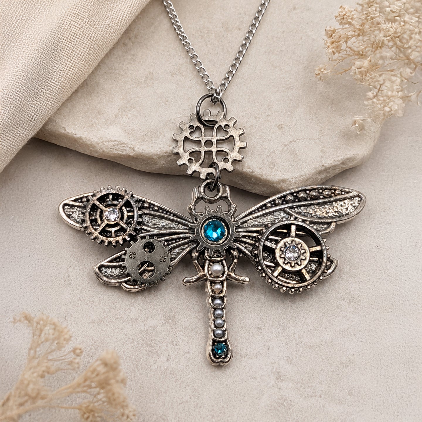 Steampunk dragonfly pendant necklace on a silver chain, featuring intricate gears, cogs, and a central blue gemstone, displayed on a textured beige stone background.