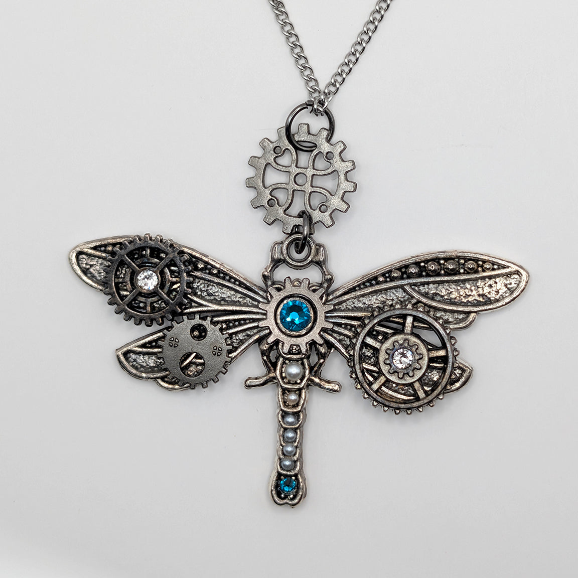 Steampunk dragonfly pendant necklace featuring intricate silver-toned gears, cogs, and wings with blue gemstone accents, hanging from a silver chain on a light gray background.
