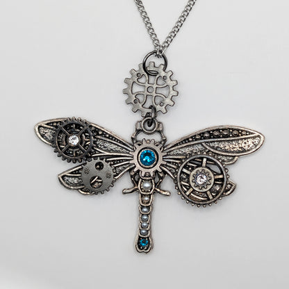 Steampunk dragonfly pendant necklace featuring intricate silver-toned gears, cogs, and wings with blue gemstone accents, hanging from a silver chain on a light gray background.