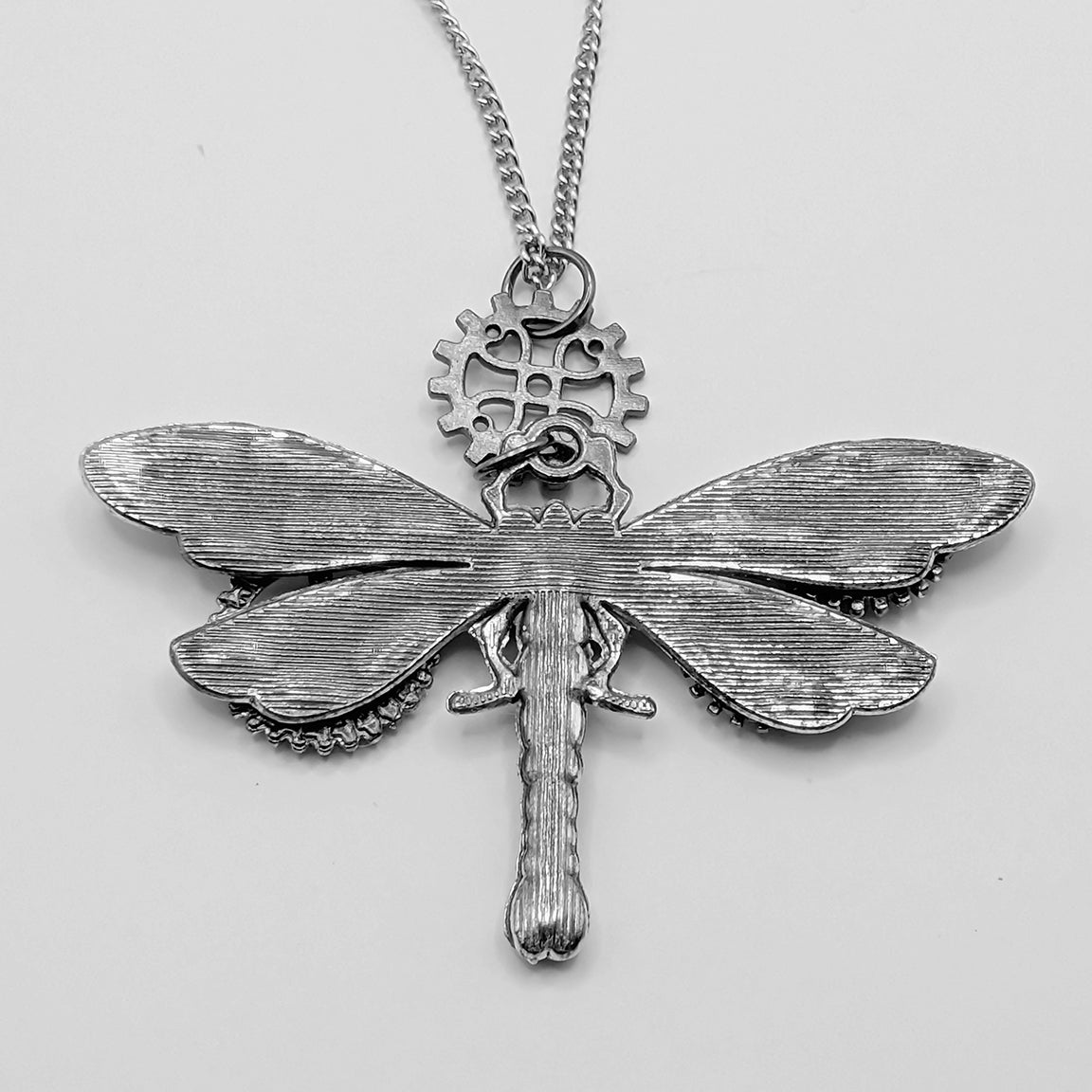 Back side of a silver dragonfly pendant necklace with intricate textured wings and a gear detail at the top, hanging from a silver chain against a light gray background.