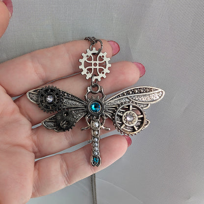Steampunk dragonfly pendant held in a hand, featuring mixed metal-toned gears, wings, and a blue crystal accent on a light gray fabric background.