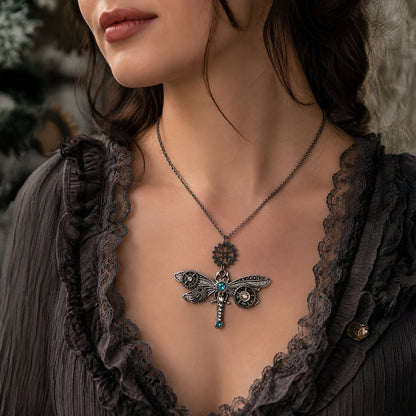 A close-up of a woman wearing a dark metal dragonfly pendant necklace with a small turquoise gemstone and clear crystal details, styled with a dark lace-trimmed top.