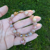 Hand holding a beaded bracelet with pink and gold accents against a green grass background