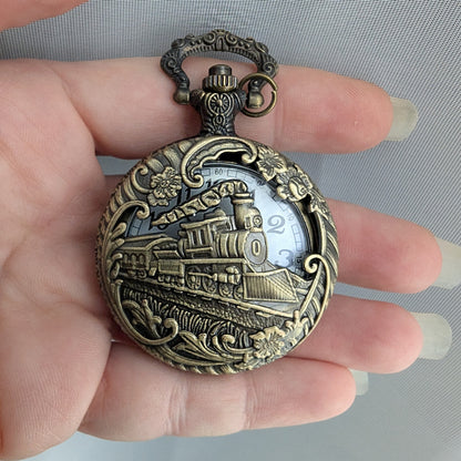 Bronze pocket watch with train design held in a hand against a gray background