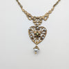Gold necklace with a heart-shaped pendant featuring pearls and a central gemstone on a light gray background.