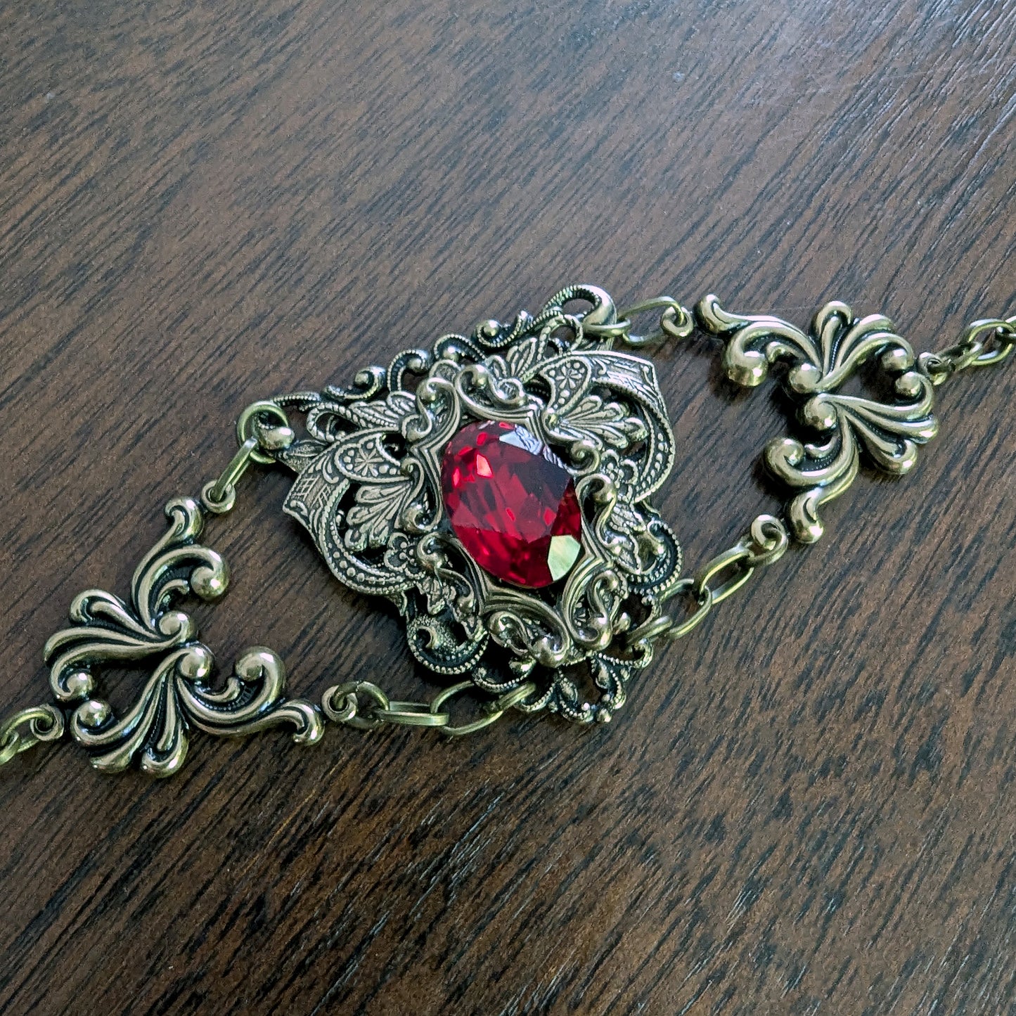 An ornate filigree bracelet with a central faceted red Swarovski crystal gemstone, set in an antique bronze-toned metal, displayed on a dark wood surface.