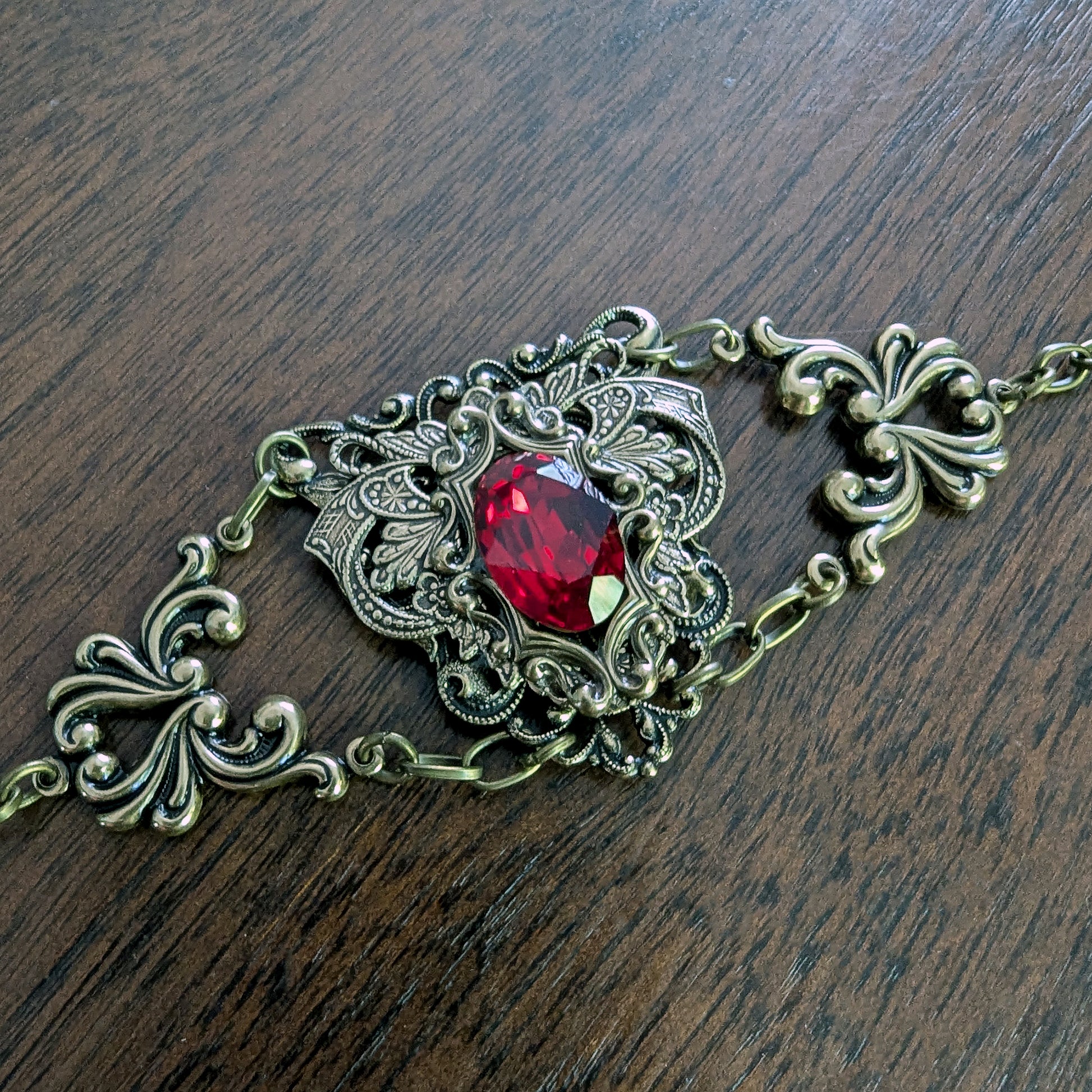An ornate filigree bracelet with a central faceted red Swarovski crystal gemstone, set in an antique bronze-toned metal, displayed on a dark wood surface.