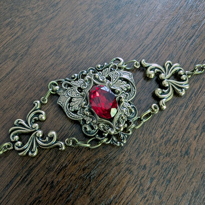 An ornate filigree bracelet with a central faceted red Swarovski crystal gemstone, set in an antique bronze-toned metal, displayed on a dark wood surface.