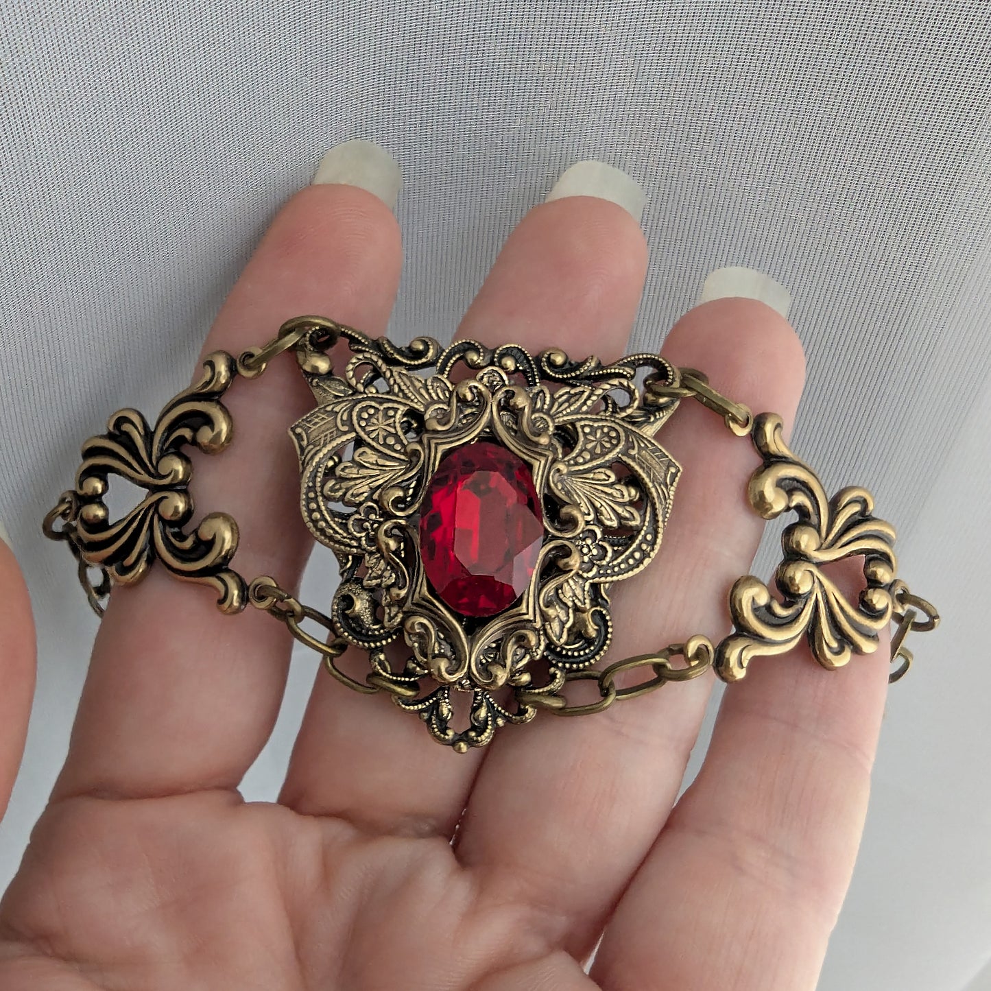 An ornate antique bronze gothic-style chain bracelet with a central faceted red Swarovski crystal gemstone, held in a hand against a light gray textured background.