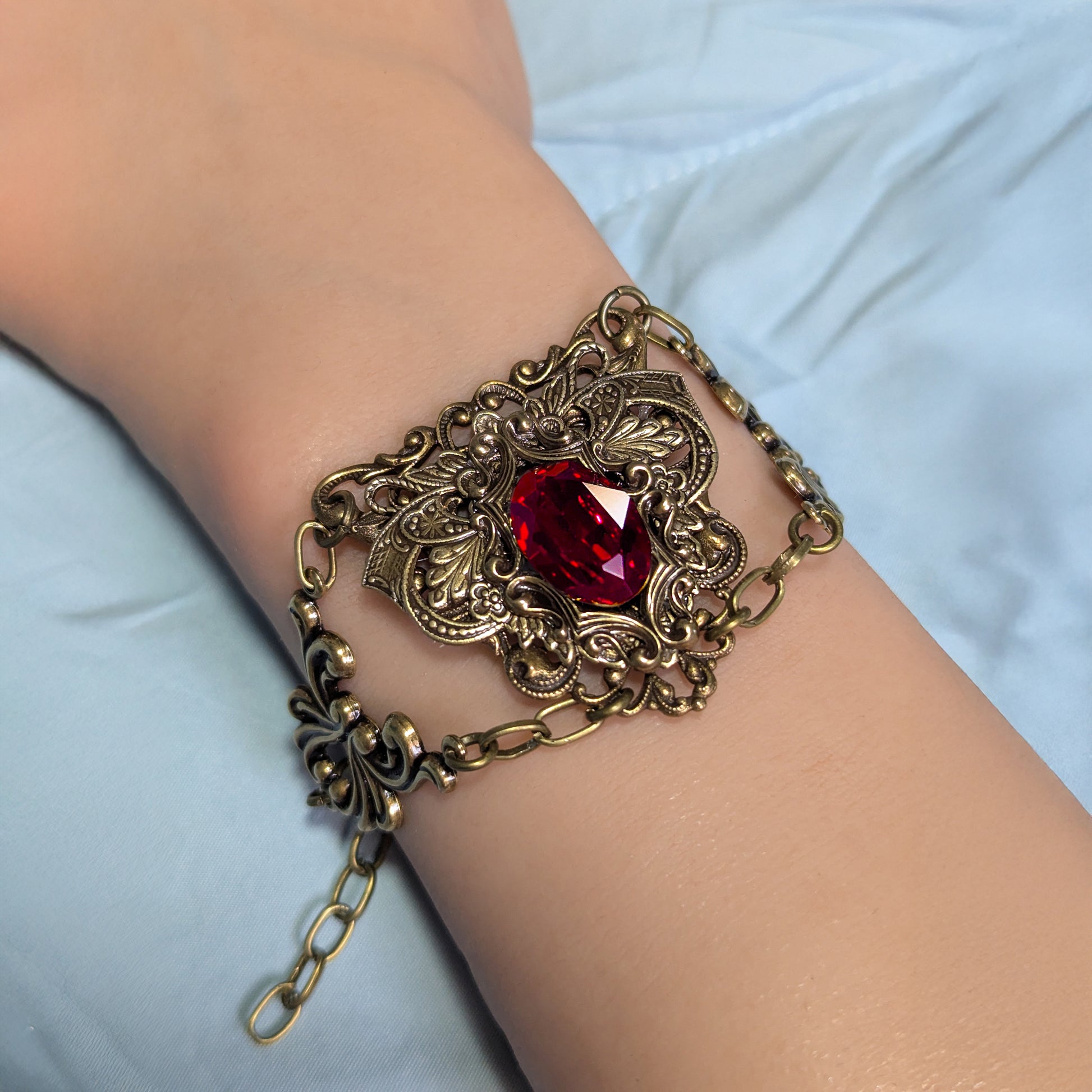 An ornate antique brass filigree bracelet with a faceted red Swarovski crystal gemstone centerpiece, shown worn on a person's wrist against a light blue fabric background.