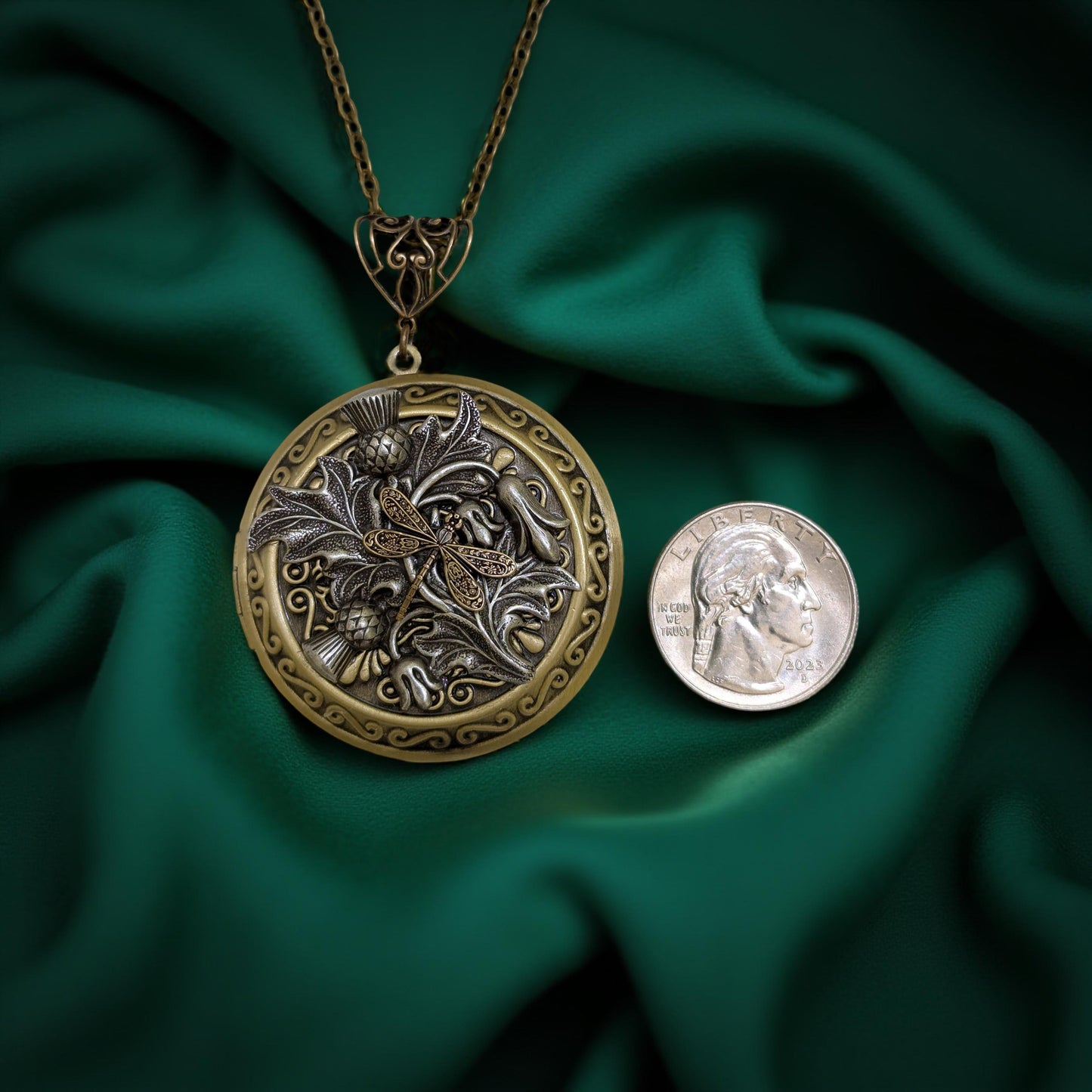 An ornate round locket pendant necklace with a peacock design, displayed next to a US quarter coin for scale on a draped dark green satin fabric background.