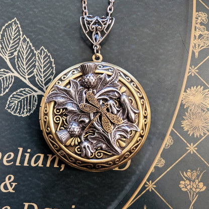 An antique-style round locket pendant necklace featuring a dragonfly motif over leaves and berries, displayed on a dark textured book page with gold botanical illustrations.