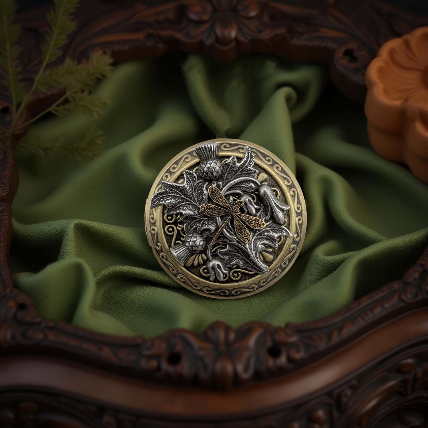 A round ornate lcoket necklace with a dragonfly motif, featuring silver-toned and gold-toned metalwork, displayed on draped olive green fabric inside an ornate dark brown wooden frame.