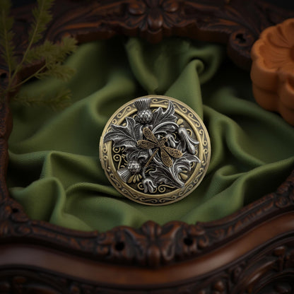 A round ornate lcoket necklace with a dragonfly motif, featuring silver-toned and gold-toned metalwork, displayed on draped olive green fabric inside an ornate dark brown wooden frame.