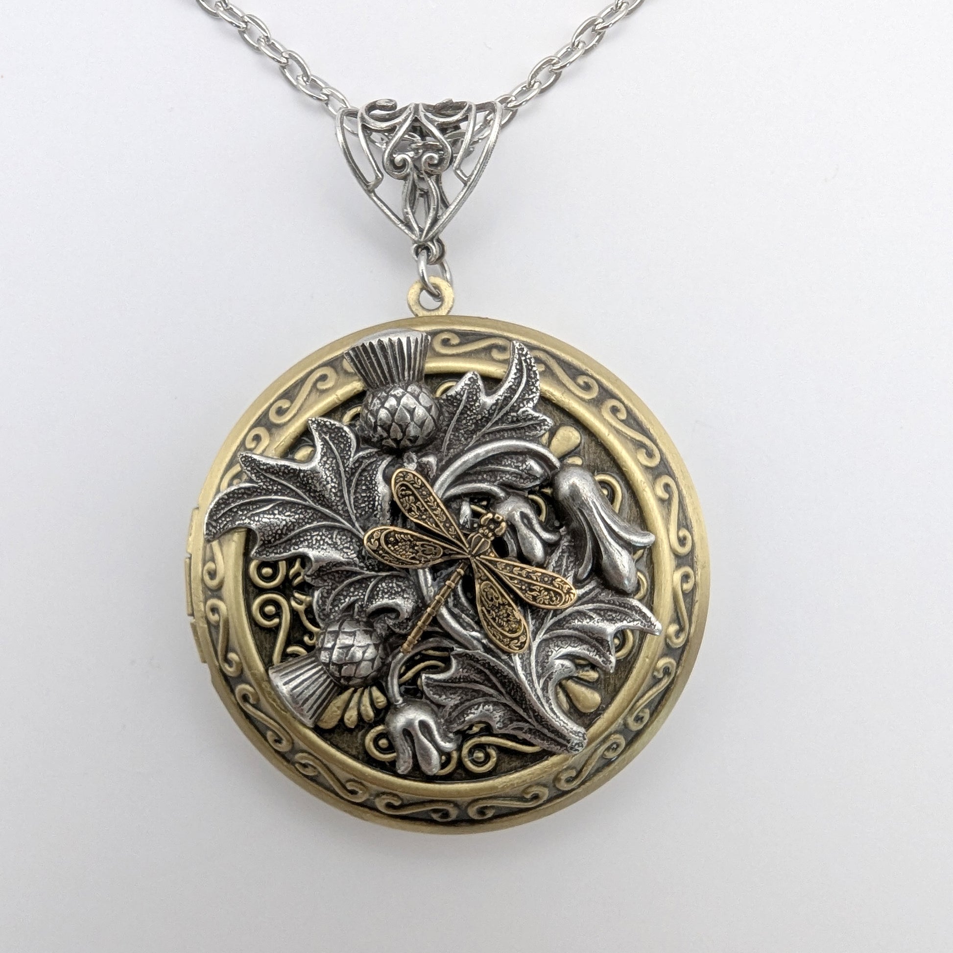 An ornate round locket pendant necklace featuring a dragonfly motif in mixed silver and gold-tone metals, suspended from a silver chain against a plain white background.