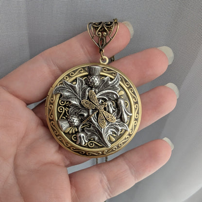 A hand with manicured nails holds a round locket pendant featuring a dragonfly and thistle design in antique gold and silver-tone metals, set against a gray textured fabric background.