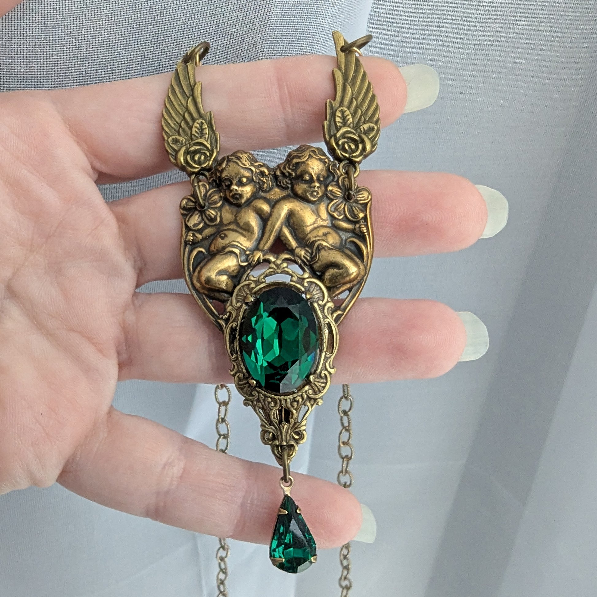 Detailed view of brass cherub pendant with emerald Swarovski crystal centerpiece and teardrop accent