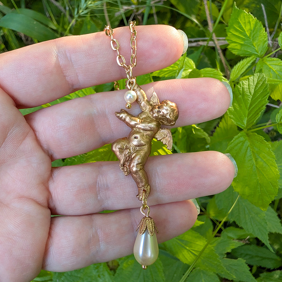 Gold cherub pendant with pearl on a chain held in a hand against a green leafy background.  Angel Jewelry