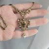 Gold heart-shaped necklace with intricate designs held in a hand against a neutral background