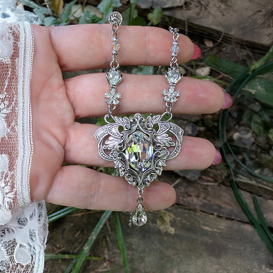 A close-up of a hand holding a sterling silver vintage styled pendant necklace with clear faceted Swarovski crystals, set against an outdoor garden background.  Fantasy bride styled jewelry.