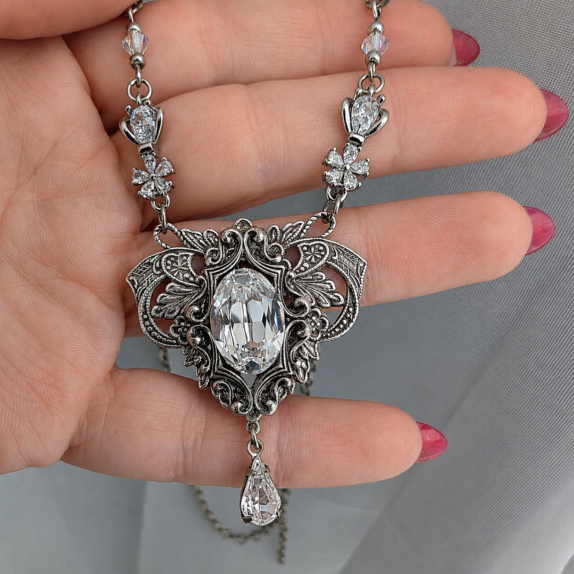 A close-up of a hand holding a sterling silver vintage styled pendant necklace with clear faceted Swarovski crystals, set against a grey fabric background.  Fantasy bride styled jewelry.