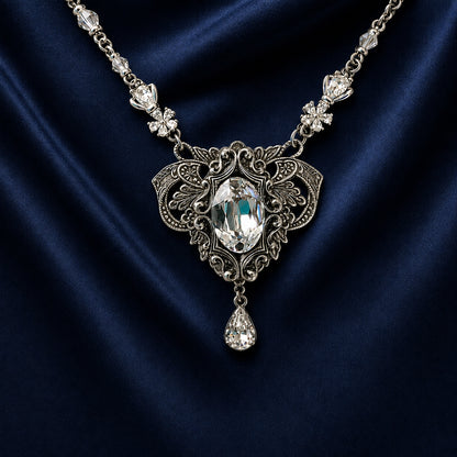 An ornate silver vintage-style necklace with a marquise-cut clear Swarovski crystal pendant and smaller clear crystals along the chain, displayed on draped dark blue satin fabric.  ELven inspired Fantasy woodland bride jewelry.