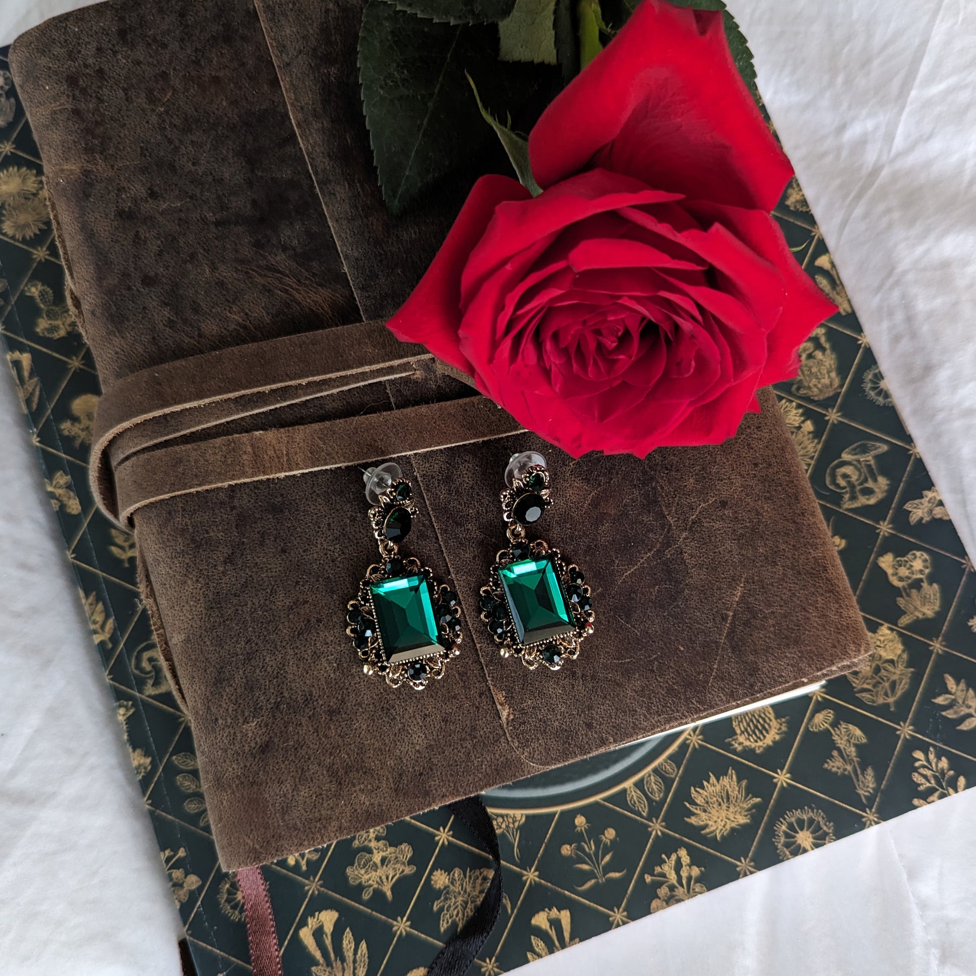 Vintage emerald green crystal earrings displayed on a brown suede book with a red rose. 