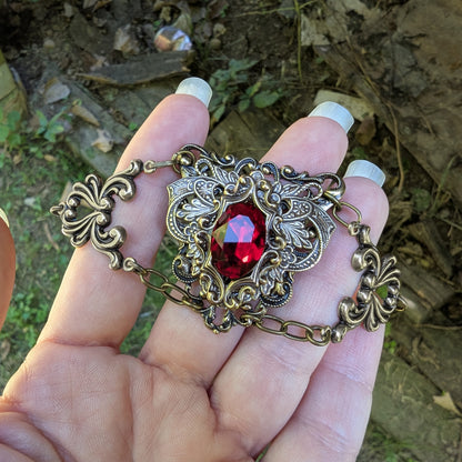 An ornate antique-style bronze chain bracelet with a central red faceted swarovski crystal gemstone and filigree details, held in a person's hand against an outdoor leafy background.