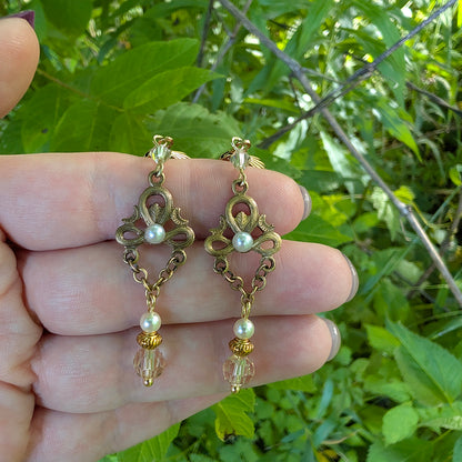 Vintage styled antique brass pearl and crystal earrings held between fingers against a green leafy background