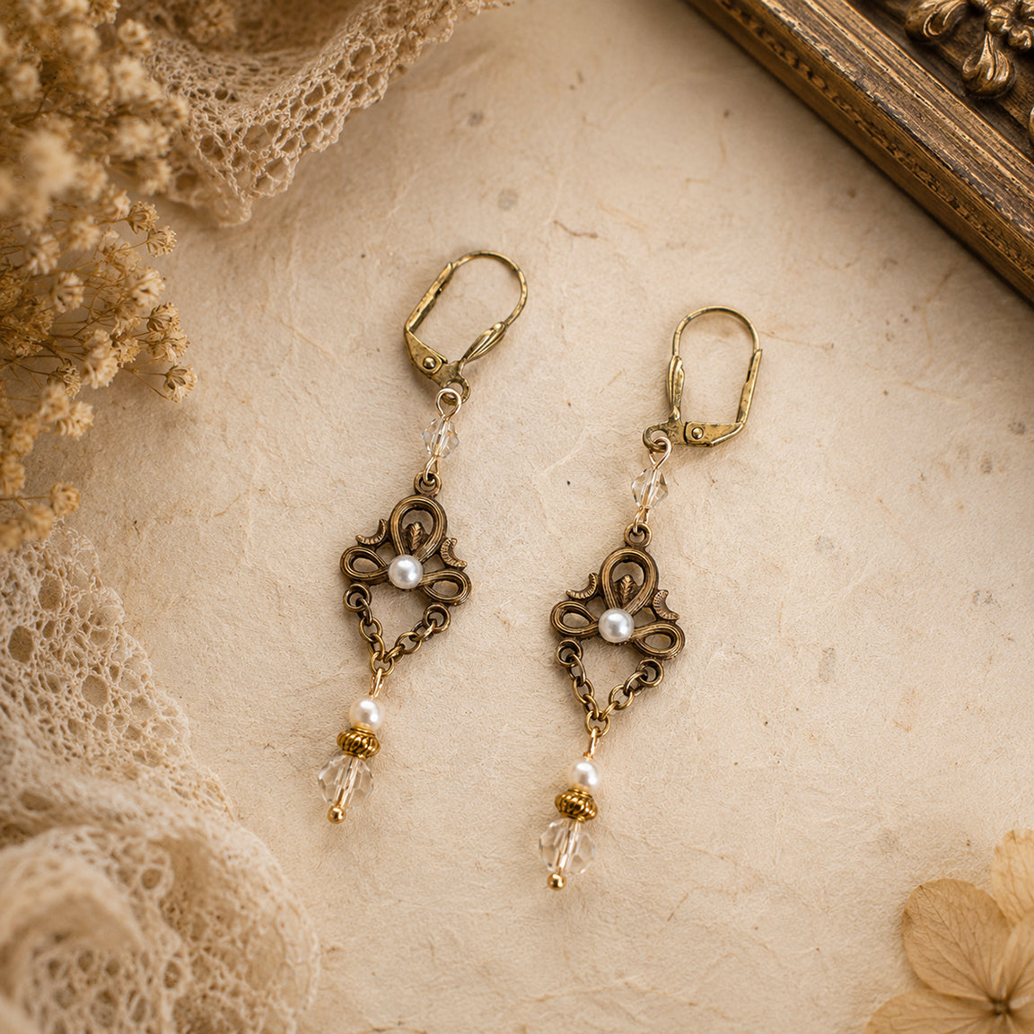 Pair of ornate vintage earrings with pearl and crystal details on a textured surface with lace and flowers.